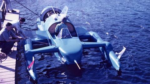 An image of Bluebird being lowered into the water at Ullswater taken in 1955. A man is inside the hydroplane, while three men stand on a pier holding ropes to lower the craft. Crowds are gathered on the edge of the water, watching on.