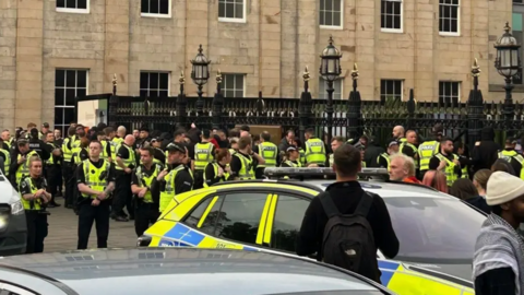 Police Scotland officers in St Andrew Square in Edinburgh. Several officers wearing high-viz vests in a fluorescent yellow colour are standing on the pavement next to black railings. A police car is visible in front of them next to a man wearing all black and a bag with the North Face logo in white.