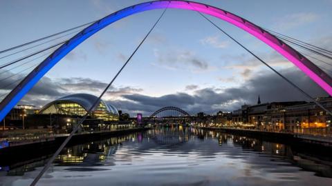 View down the River Tyne at dusk through the arch of the Millennium Bridge, which is illuminated in blue and purple, looking down towards the Tyne Bridge. On either side the buildings are lit up, with reflections in the water.