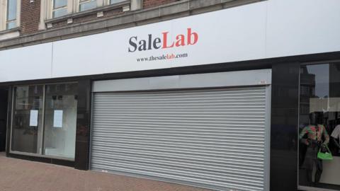 A shop front with shutters down. There is white sign with red and red lettering that read Sale Lab.