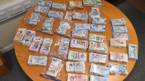 Stacks of money wrapped held together by elastic bands, some placed inside sandwich bags, sit on a wooden table. There is about twenty of them. 