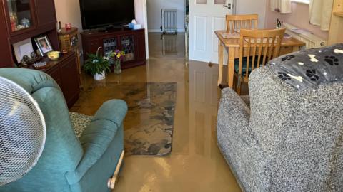 The inside of a lounge, filled with water, there is dirty water on the ground. There are two chairs, a table, chairs, a carpet and furniture. 