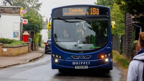 A blue single-decker bus with the destination Cambridge via Cambourne and Comberton written on a digital display above the windscreen. It is facing the camera on a narrow village road, with a post office and post box on the side of the road. 