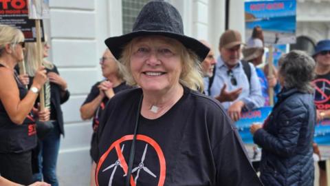 Georgina is wearing a black hat and black t-shirt with two turbines on it with a circular symbol with a line across it over them. She has shoulder length fair hair and is standing in front of protestors with placards by a white building.