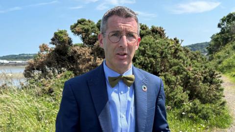 A man with glasses stands outside. He is wearing a navy suit, blue shirt and green dickie bow He is standing in front of a bush. It's a bright day, the sky can be seen with a couple of faint clouds.