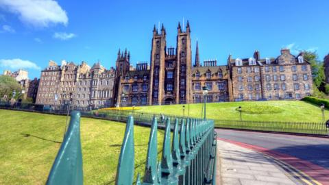 A view of Edinburgh University's New College buildings on a sunny day - there are blue skies and green grass around it, while the buildings themselves include twin towers. 