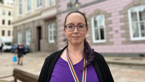 A dark haired woman with glasses in purple top and black cardigan stands in front of of a granite building with pink tiles on the outside.
