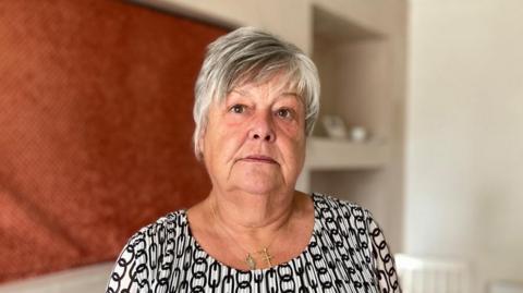 A woman with short white hair wearing a black and white top and gold jewellery stands in a white and orange house. 