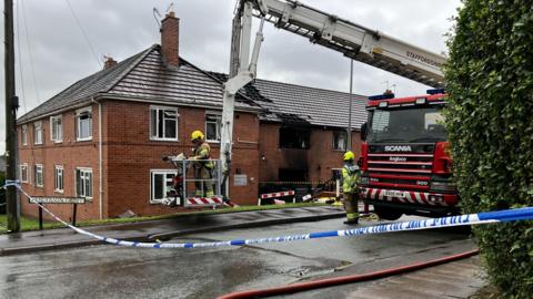 Police tape is cordoning off a road. There is a fire engine with an aerial platform parked next to a block of two-storey flats. A section of the flats is black with smoke damage and there are roof tiles missing. There are two fire officers, one in the aerial lift, and one standing by the fire engine