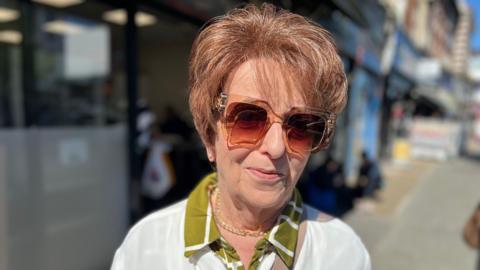 Woman in white jumper, green shift and big sunglasses looks at camera outside a shop