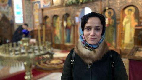 A woman with a black coat, fur round the neck and a blue headscarf stands in the church, although the background is blurred. She has dark hair and is smiling. 