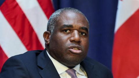 David Lammy looks ahead while sitting down in a dark blue suit and a patterned tie made up of small white and pink checks. The flags of the US and UK can be seen behind him.
