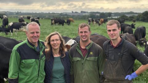 A family of four stand together in a field with diary cows in the background. They are wearing work wear or overalls. One is also wearing blue plastic gloves that are used in a diary parlour. 
