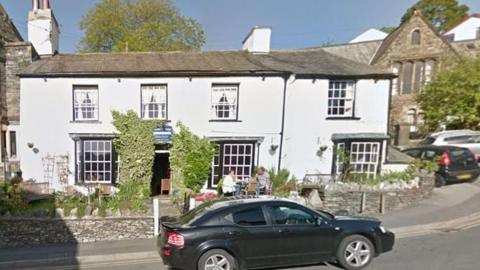 A Google Maps screenshot of the outside of Laurel Cottage in Bowness. It is painted white with black window frames and has ivy around its open door. There is a couple sitting at a table outside. 