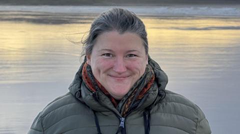Lucy Cowley yn sefyll o flaen llyn. Mae hi'n gwisgo cot werdd ac edrych tuag at y camera.