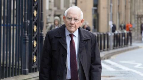 Sir Duncan Nichol, who has short white hair, wearing glasses and a white shirt with a burgundy tie under a black coat, walks towards Liverpool Town Hall. 