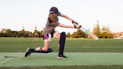 A girl bats wearing a helmet, shin pads and gloves. She is on a cricket pitch in the early evening