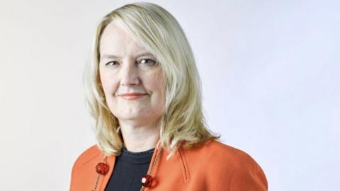 A headshot of Prof Helena Teede wearing a black top with an orange jacket and a necklace with orange beads on it. She has shoulder length blonde hair and she is smiling at the camera.