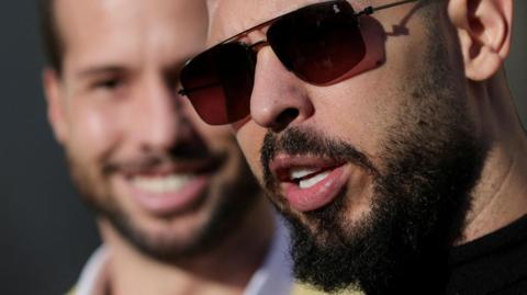 A head shot of Andrew Tate up close and just out of focus behind him is his brother Tristan. Sunshine is lighting their faces. Andrew is wearing sunglasses and has a black curly beard and Tristan has a similar beard. Andrew appears to be talking while Tristan is smiling. 