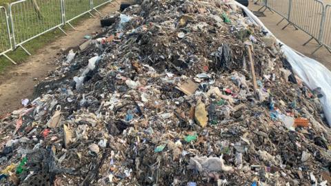 A huge pile of waste dumped in a car park at a park. It has been closed off by metal fencing. The car park has a mud surface and is surrounded by wooden posts and grass.