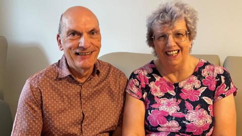 An elderly man with a brown shirt smiles as he sits on a sofa next to an elderly woman wearing a black and pink floral top, who is also smiling.