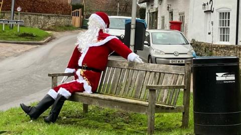 A model of Santa Claus sat on a bench on a village green.