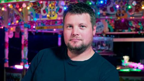 Raecher with short hair and short beard looks at camera in front of ferris wheel