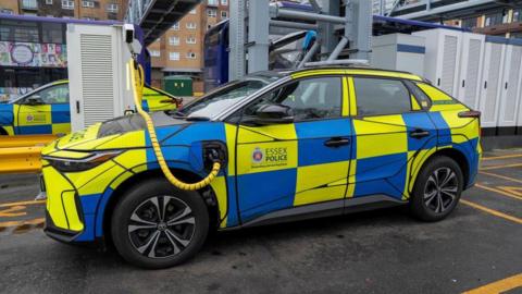 An Essex Police car charges up at an electric vehicle charging station at the First Bus depot in Basildon. The car is covered in blue and yellow squares, and there is a yellow and black charging cable reaching from a terminal across the bonnet of the vehicle and into its left-hand side.
