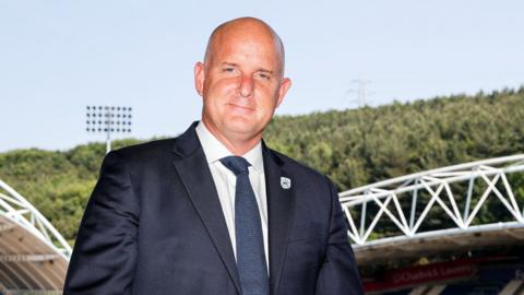 Jake Edwards stands against the backdrop of The Accu Stadium, with blue sky and trees in the background. he has a small closed mouth smile and wears a navy suit, dark blue tie and white shirt