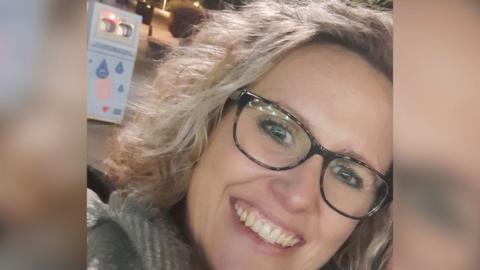 Picture of Kaeti Morrison smiling in a selfie. She has blonde wavey hair and is wearing glasses. Behind her is a small grey machine with a red light. 