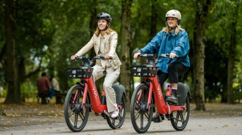 Two women ride red electric Voi bikes through a leafy park. 
