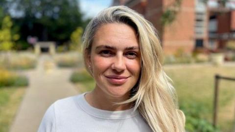 Jenna Robinson is smiling into the camera. She has long blonde hair and is wearing a white top. She is standing in a public garden which has wild flowers in it behind a council building in Beverley.