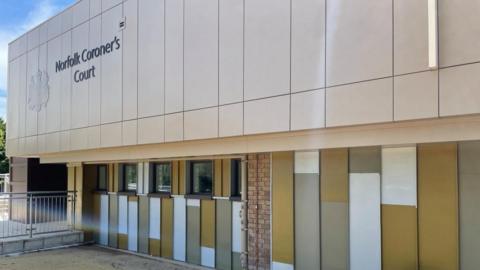 The Norfolk Coroner's Court is a building clad with white, grey and mustard coloured panels, with brown windows. There is a royal crest and the words "Norfolk Coroner's Court" beneath it. There are railings towards the left side of the picture.