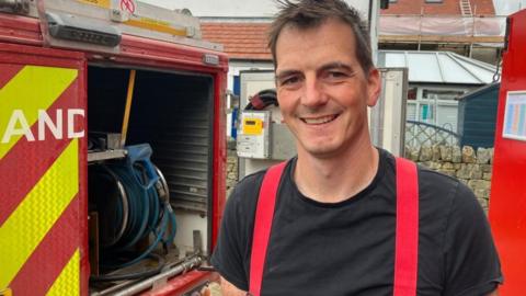 A man with short brown hair looks into the camera and smiles. He is wearing a black T-shirt with red braces holding up firefighter uniform trousers. He is standing in front of the back door of a firefighting Land Rover, which has the back door open showing a blue hose.