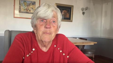 Blockley seated indoors at a table, facing the camera directly. She has short, neatly cut white hair and light-colored eyes, with natural facial lines visible around the eyes, cheeks, and mouth. She is wearing a red knit sweater that features decorative metallic buttons along the shoulder seams, and the forearms are resting on a surface just out of frame, suggesting a seated, slightly leaning-forward posture.