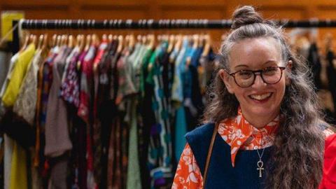 A woman in vintage clothing from the seventies and wearing glasses, smiling in front of a rack of clothes