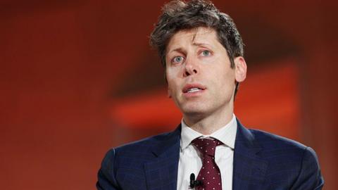 Sam Altman against red background, wearing blue suit and red tie with white dots, looks up and to the side
