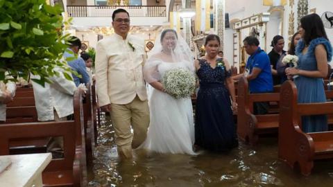 Bride wades through flooded church. She wears white and holds a large bouquet of baby's breath