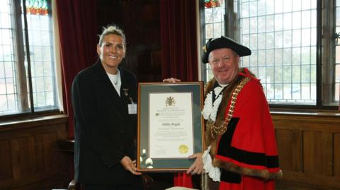 A woman with a black suit and tied-back blonde hair holds an award with a man in a traditional, red ceremonial outfit and hat. 