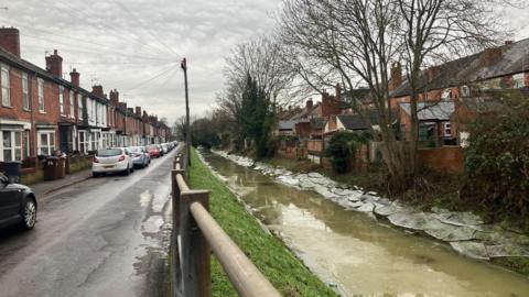 A street with terrace houses on the left and cars parked in single file. A fence in the middle is a barrier between the road and the dyke. The dyke on the right has brown water and remnants of ice