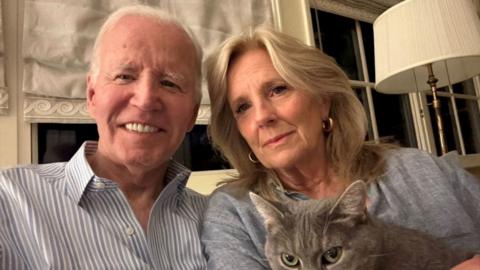 Joe Biden and Jill Biden pose for a selfie photo. They are sat down in front of a curtained window, Jill is holding a grey cat.