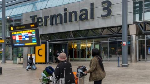 This image captures the exterior of Terminal 3 at an airport. The terminal's name is displayed prominently in large black letters on the building. In the foreground, an information board showcases flight details, complete with airline logos. Two individuals stand with luggage carts, scanning the board for information. The surrounding scene includes other travellers and the entrance to the terminal, marked with a large yellow "C" sign