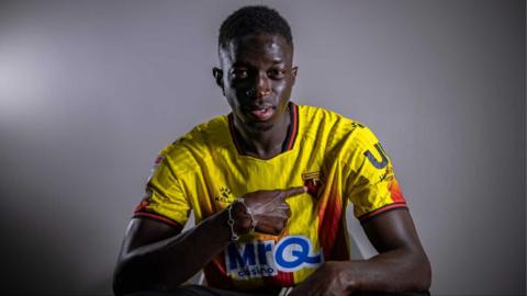 Formose Mendy looks straight ahead with a wry smile as he points to the Watford badge on his yellow and red stripe kit.