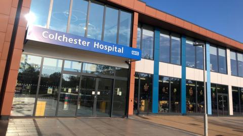 The entrance to Colchester Hospital, which is a two-storey building with a glass facade. It has a blue banner that says Colchester Hospital above the entrance doors.