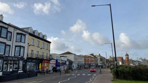 Street view picture of Morecambe. A row of shops are on the left of the road, whilst a grassy verge is on the right. A car drives past the camera and it's a blue sky.