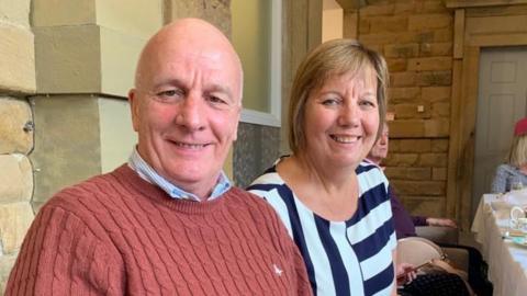 A middle-aged couple posing for a picture during dinner with a larger group of people. The man, on the left, is wearing an orange jumper with a blue shirt underneath. The woman, on the right, is wearing a navy and white striped top