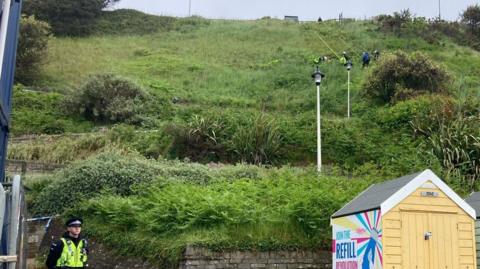 A group of officers abseiling down the green sloping cliffs while a uniformed officer stands guard at the bottom