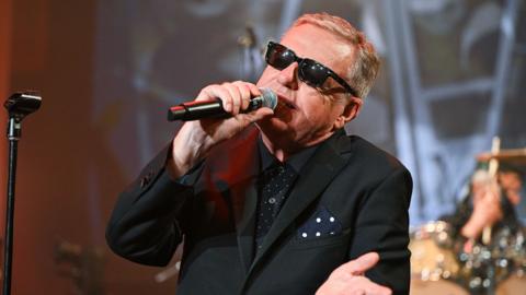 Madness performs in the BBC Radio Theatre. Lead singer Suggs is wearing a black suit and sunglasses as he sings into a microphone