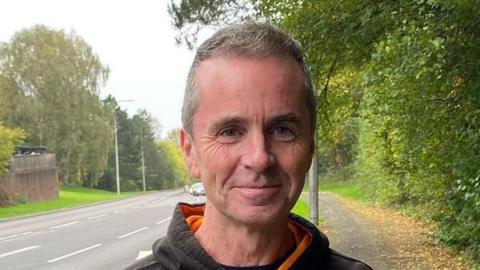 A photograph of a man's face with a road behind him. Trees and lampposts can be seen either side of him.