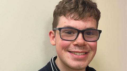 Close-up of young man's face. He is wearing black glasses and is smiling.